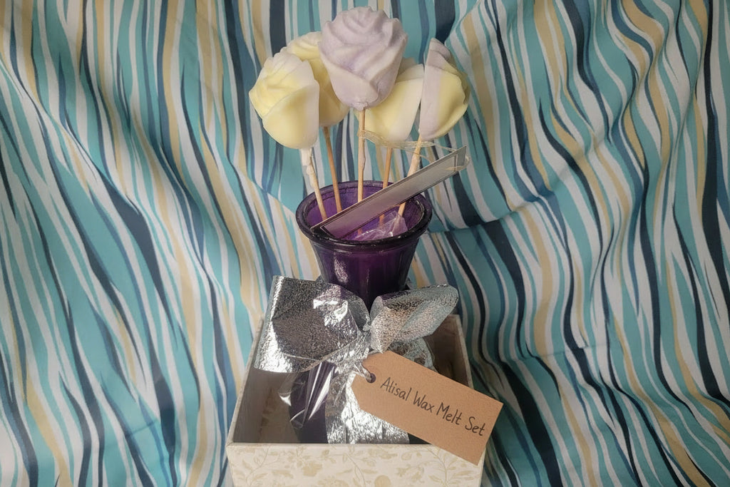 Decorative wax melts including a silver bow, purple container, and skewer with yellow and white decorative elements on a striped fabric background.