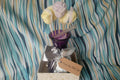 Decorative wax melts including a silver bow, purple container, and skewer with yellow and white decorative elements on a striped fabric background.