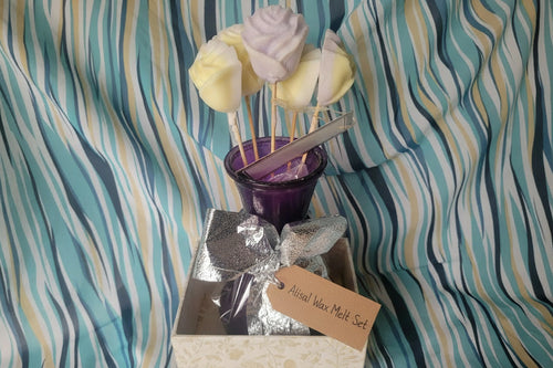 Decorative wax melts including a silver bow, purple container, and skewer with yellow and white decorative elements on a striped fabric background.